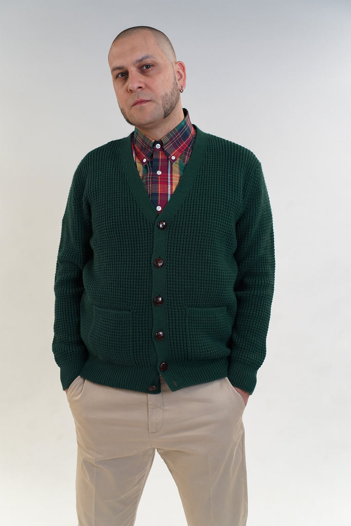 Man wearing a green cardigan over a plaid shirt with beige pants on a white background