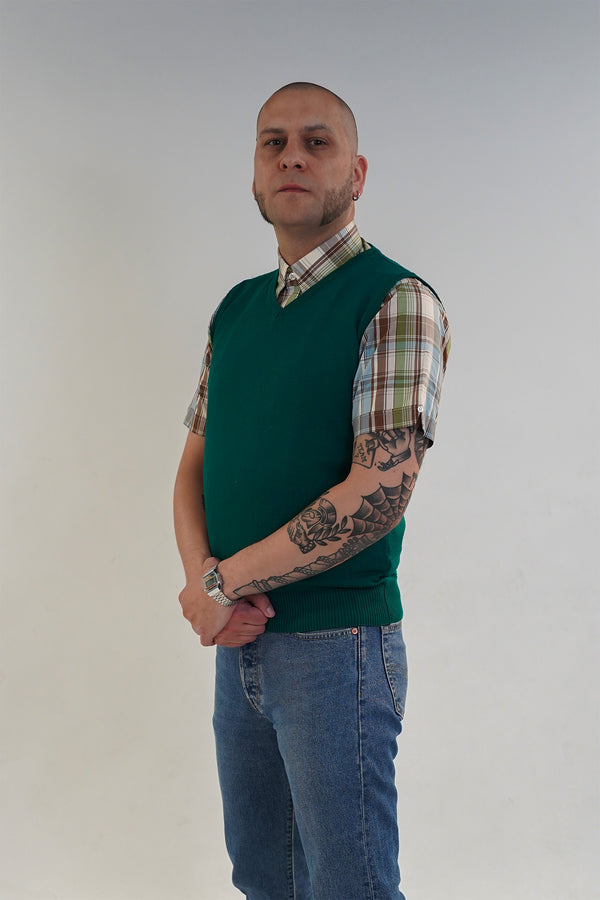 Man wearing a green sweater vest over a plaid shirt with rolled-up sleeves, standing against a plain background.
