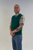 Man wearing a green sweater vest over a plaid shirt with rolled-up sleeves, standing against a plain background.