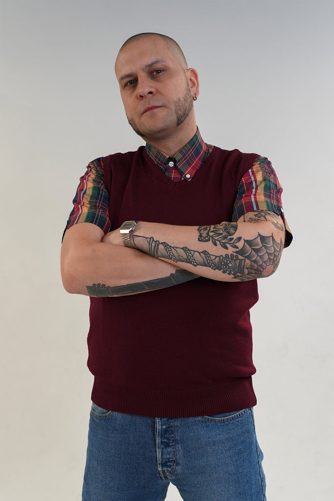 Man wearing a maroon sweater with plaid sleeves and blue jeans against a plain background