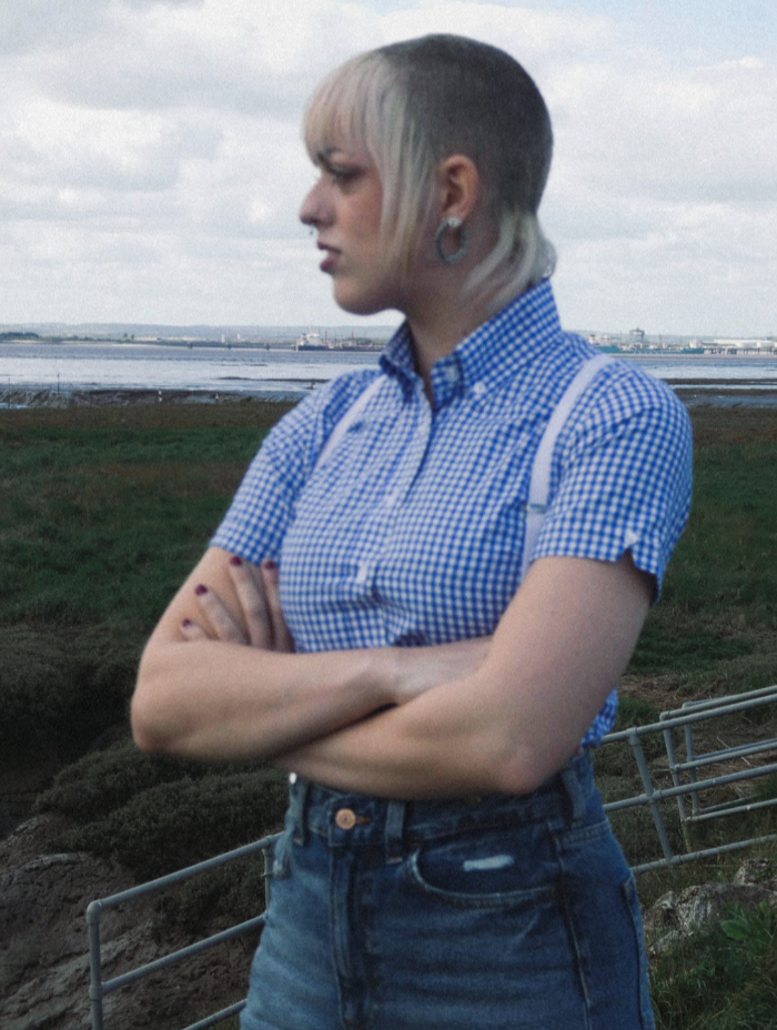 Instagram legend deadbeatd0lly wearing a blue gingham checkered shirt and white braces standing outdoors with a scenic background.